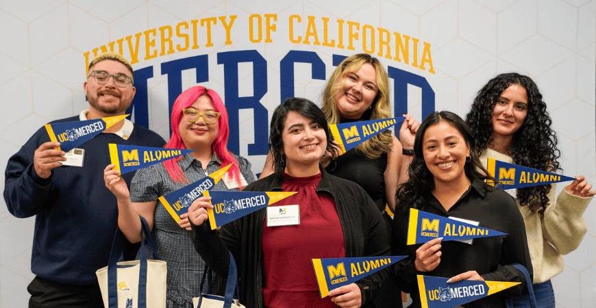 Six UC Merced students pose in front of a University of California, Merced backdrop, smiling and holding blue-and-gold pennants that read “UC Merced Alumni” and “UC Merced.”